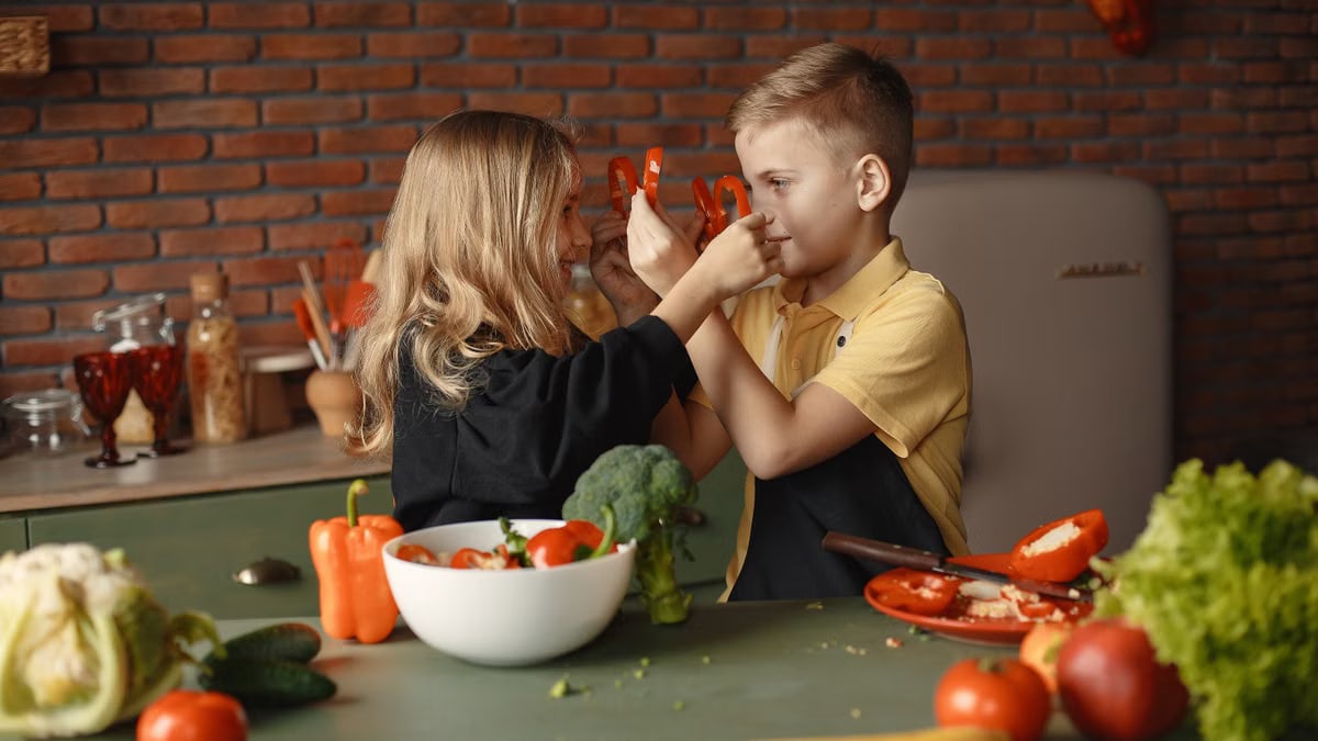 Playful boy and girl wearing apron preparing healthy vegetarian salad together at table with ingredients and bowls while looking at each other through slices of pepper